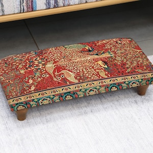 May include: A rectangular footstool with a detailed, patterned fabric top. The fabric features a red background with a design of birds and floral elements in shades of gold, green, and blue. The footstool is supported by four short, wooden legs.