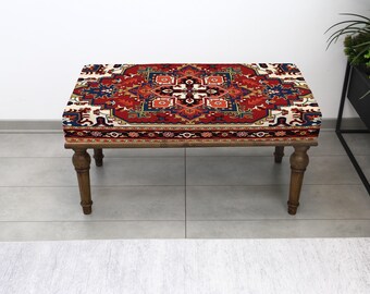 Puede incluir: Banco rectangular de madera con tapa estampada. La tapa presenta un diseño rojo, azul y crema que recuerda a una alfombra tradicional. El banco tiene cuatro patas de madera torneada, apto para uso en interiores.