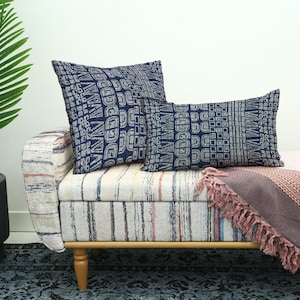 May include: Two navy blue throw pillows with a geometric pattern rest on a striped bench. A pink blanket with fringe is draped over the bench. A potted palm tree is to the left.