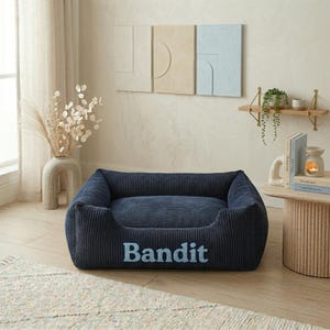 May include: A navy blue corduroy dog bed with the name "Bandit" embroidered in light blue. The bed has a rectangular shape with raised sides and a plush interior. The bed is designed for pets.