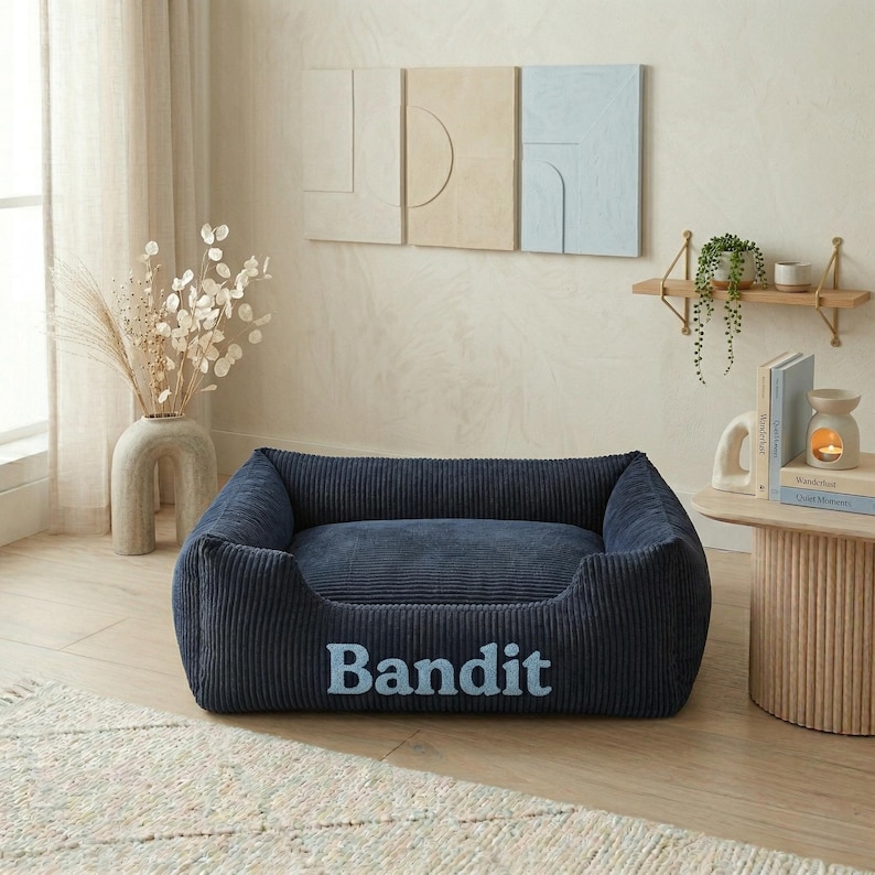 May include: A navy blue corduroy dog bed with the name "Bandit" embroidered in light blue. The bed has a rectangular shape with raised sides and a plush interior. The bed is designed for pets.