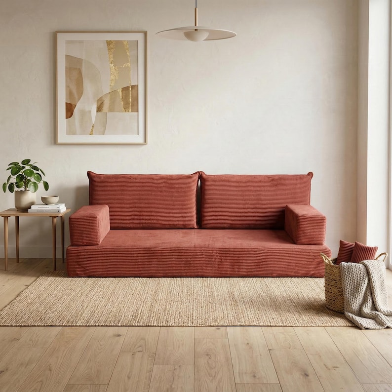 Op de afbeelding: Een terracotta corduroy bank met twee rugkussens en armleuningen staat op een geweven jute vloerkleed. Een abstracte ingelijste kunstwerk hangt aan de muur. Een kleine houten bijzettafel bevat een plant en boeken.