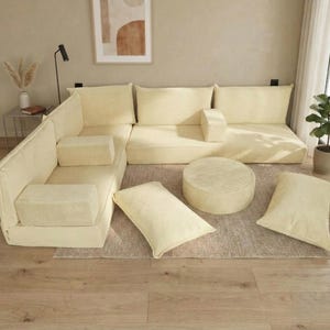 Op de afbeelding: Een modulaire hoekbank in cr&egrave;mekleurig corduroy. De set bevat een hoekstuk, armloze secties, een ronde poef en diverse rechthoekige en vierkante kussens. De meubels zijn geplaatst op een neutraal getint tapijt.