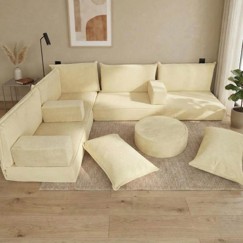 Op de afbeelding: Een modulaire hoekbank in cr&egrave;mekleurig corduroy. De set bevat een hoekstuk, armloze secties, een ronde poef en diverse rechthoekige en vierkante kussens. De meubels zijn geplaatst op een neutraal getint tapijt.