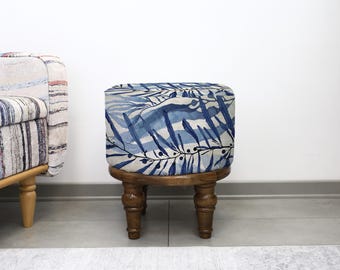 May include: A small, square footstool with a wooden base and a blue and white leaf pattern on the cushioned top. The footstool has four turned legs and is placed in front of a white wall.