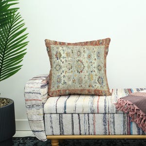 Pode incluir: Uma almofada decorativa com um design de tapete vintage. A almofada apresenta um fundo creme com um padrão detalhado em tons de vermelho, azul e amarelo. Está colocada num banco listrado, com uma manta com franjas. Mede cerca de 40 cm x 40 cm.