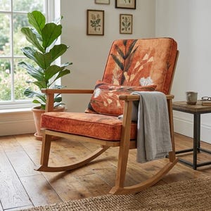 May include: Wooden rocking chair with orange floral upholstery and a matching cushion. A grey blanket is draped over the arm. A small side table with a mug and glasses is next to the chair. A large plant is in the background.