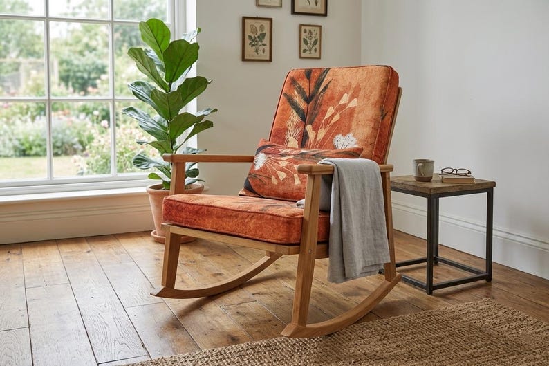 May include: Wooden rocking chair with orange floral upholstery and a matching cushion. A grey blanket is draped over the arm. A small side table with a mug and glasses is next to the chair. A large plant is in the background.