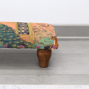 May include: A decorative footstool with a colorful fabric featuring a peacock, deer, and lotus flowers. The fabric is stretched over a rectangular frame and supported by four wooden legs. The color palette includes shades of orange, blue, and green.