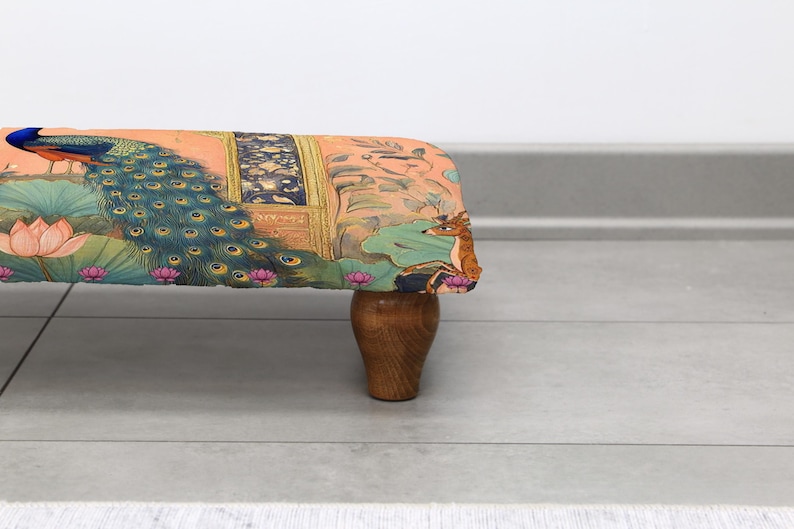 May include: A decorative footstool with a colorful fabric featuring a peacock, deer, and lotus flowers. The fabric is stretched over a rectangular frame and supported by four wooden legs. The color palette includes shades of orange, blue, and green.