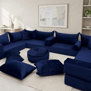 May include: A modular navy blue velvet sectional sofa with multiple cushions and a round ottoman. The sofa is arranged in a U-shape on a cream-colored rug, creating a comfortable seating area. Decorative pillows add to the cozy aesthetic.