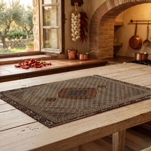 May include: A rectangular rug with a central diamond design in shades of brown, beige, and rust. The rug is placed on a rustic wooden table, showcasing a detailed, geometric pattern. The rug's border features a repeating floral motif.