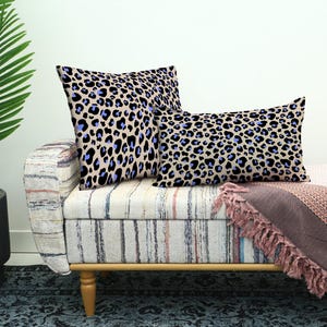 May include: Two decorative throw pillows with a beige leopard print pattern featuring black and blue spots. The pillows are placed on a striped bench with a pink blanket and a potted plant in the background.