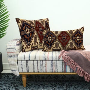 May include: Two decorative throw pillows with a geometric pattern in beige, navy, and burgundy rest on a striped bench. A matching patterned blanket with fringe is draped over the bench. A potted palm tree is in the background.