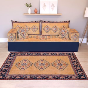 May include: A living room set with a sofa and rug, both displaying matching geometric patterns in gold, blue, and red. The sofa has a navy blue base and decorative cushions. A wooden floor lamp and wall art complete the scene.