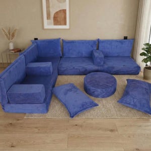 May include: A modular blue corduroy sectional sofa with multiple cushions and a round ottoman. The sofa is arranged in an L-shape, with additional cushions and a round ottoman. The furniture is placed on a neutral-colored rug.