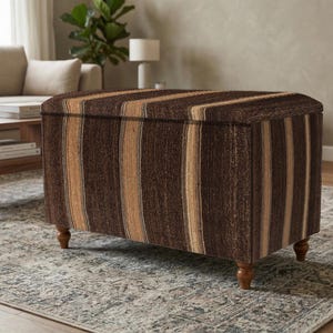 May include: A rectangular storage ottoman with a striped pattern in shades of brown and tan. The ottoman has a hinged lid and sits on four short, turned wooden legs. It is placed on a patterned rug.