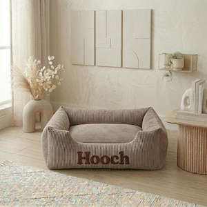 May include: A corduroy dog bed in a neutral beige color. The bed has a rectangular shape with raised sides and a plush interior. The name "Hooch" is embroidered in a darker brown font on the front of the bed.