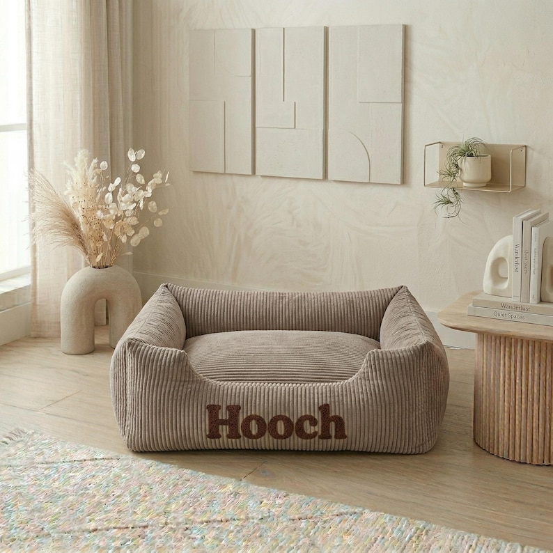 May include: A corduroy dog bed in a neutral beige color. The bed has a rectangular shape with raised sides and a plush interior. The name "Hooch" is embroidered in a darker brown font on the front of the bed.