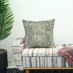 May include: A square cushion with a faded, vintage-style pattern in grey, green, and red hues. The cushion sits on a striped bench. A potted palm tree is visible on the left. The cushion's pattern resembles a rug.