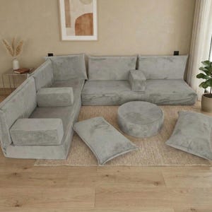 Może przedstawiać: Modułowa sofa narożna z jasnoszarego sztruksu. Zestaw zawiera elementy narożne, oparcia, podłokietniki, okrągłą pufę i kilka prostokątnych poduszek. Meble ustawione są na dywanie w neutralnych odcieniach, tworząc przytulną przestrzeń mieszkalną.