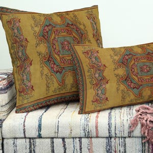 May include: Two decorative pillows with a mustard yellow background and a floral design in shades of pink, teal, and red. The pillows are rectangular and have a patterned border. They are placed on a striped sofa.