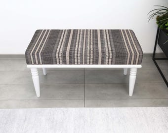 May include: A rectangular bench with a woven, striped top in shades of grey, beige, and white. The bench has a white wooden frame and four turned legs. The design is simple and modern, suitable for various interior styles.