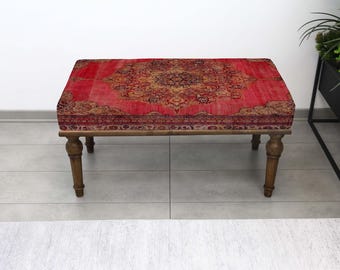 Puede incluir: Un otomán rectangular con una parte superior roja estampada y patas de madera. La parte superior presenta un diseño de medallón central en tonos dorados y marrones, rodeado por un borde. Las patas son de color marrón oscuro.