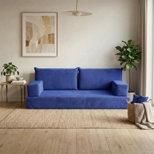 Op de afbeelding: Een blauwe corduroy bank met twee rugkussens en armleuningen staat op een natuurvezel vloerkleed. Een kleine houten bijzettafel met een plant en kunstwerk is zichtbaar. Een gevlochten mand met blauwe kussens en een deken staat rechts.