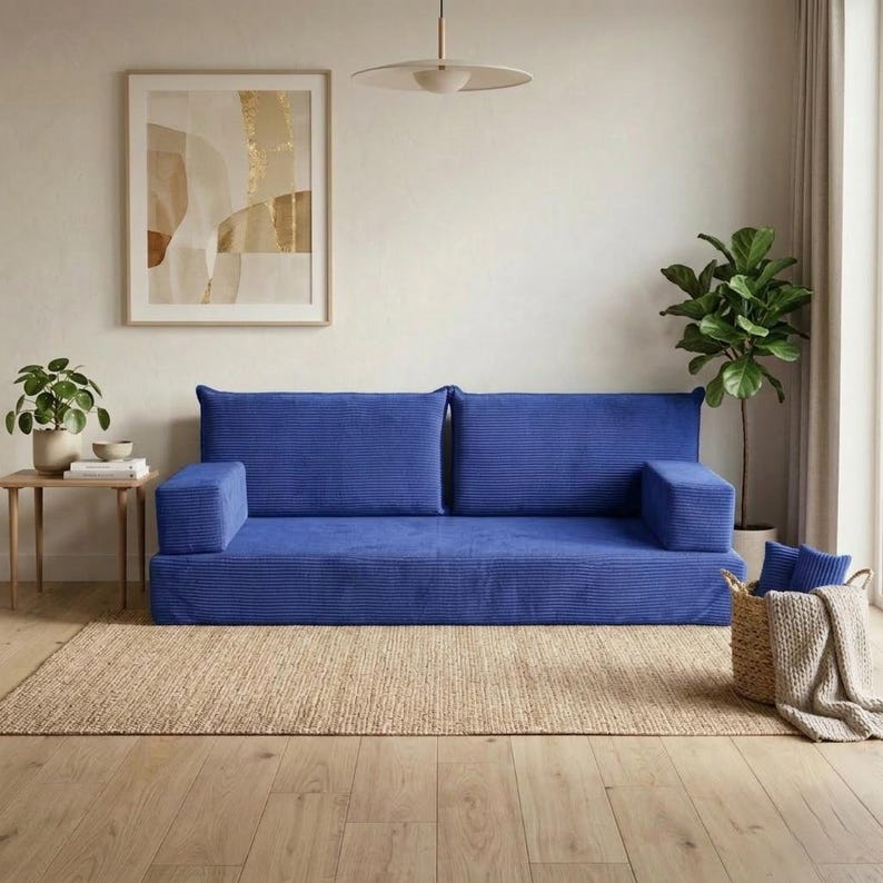 Op de afbeelding: Een blauwe corduroy bank met twee rugkussens en armleuningen staat op een natuurvezel vloerkleed. Een kleine houten bijzettafel met een plant en kunstwerk is zichtbaar. Een gevlochten mand met blauwe kussens en een deken staat rechts.