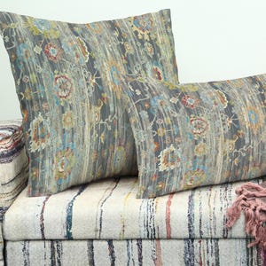 May include: Two decorative cushions with a grey, blue, and orange floral pattern rest on a patterned sofa. The cushions have a distressed, vintage look. A fringed throw blanket is draped over the sofa.
