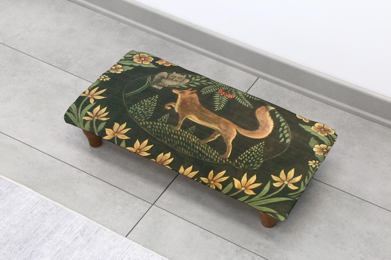 Op de afbeelding: Een rechthoekige voetenbank met een donkergroene stoffen bovenkant met een vosillustratie. Het ontwerp bevat gele bloemen en bladeren. De kruk heeft vier korte, houten poten en is geschikt voor woondecoratie.