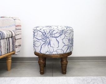 May include: A small, round footstool featuring a blue and white floral pattern. It has a dark brown wooden base with four turned legs. The design evokes a botanical theme. The dimensions are approximately 40 cm in diameter.