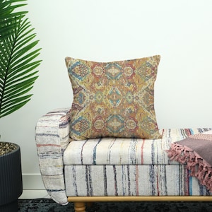 May include: A decorative square pillow with a colorful, intricate pattern in shades of gold, red, and blue. The pillow is placed on a striped bench with a wooden frame, next to a potted plant.