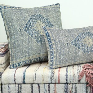 May include: Two decorative throw cushions with a blue and grey patterned design rest on a patterned sofa. The cushions feature a central diamond motif and border details. A fringed throw blanket is visible.