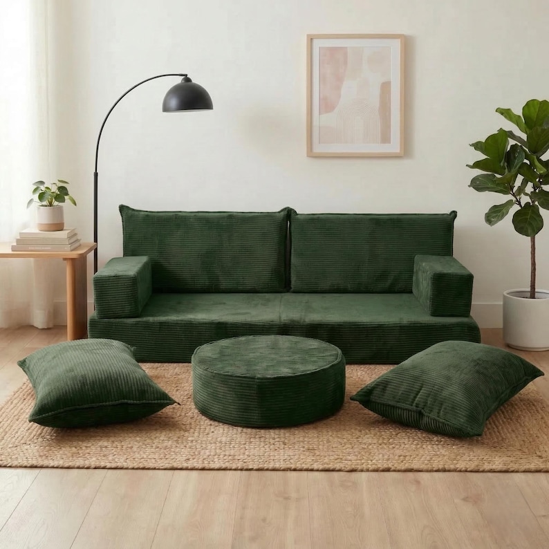 Op de afbeelding: Een modulaire groene corduroy bankstel met een bijpassende ronde poef en twee vierkante kussens. De set staat op een geweven jute vloerkleed, wat een gezellige en uitnodigende woonruimte cre&euml;ert. Een vloerlamp en potplanten maken de kamer compleet.