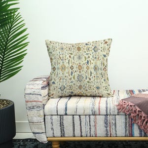 Pode incluir: Uma almofada decorativa quadrada com fundo creme e um padrão colorido detalhado. A almofada é colocada em um banco listrado com moldura de madeira, ao lado de uma planta em vaso.
