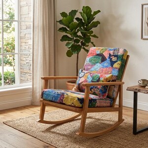May include: A wooden rocking chair with a colourful patchwork cushion and backrest. The chair sits on a woven rug in a room with a side table, a window, and framed botanical prints.