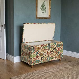 Puede incluir: Arcón de almacenamiento abierto con exterior de tela estampada. La tela presenta un diseño repetido en rojo, amarillo, azul y verde sobre un fondo crema. El interior es de color crema y descansa sobre patas cortas de madera. Mide unos 90 cm de largo.