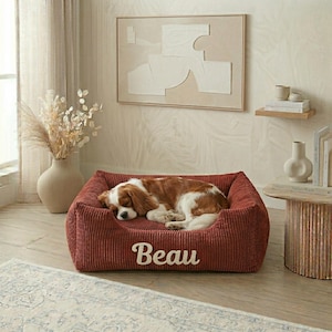 May include: A red corduroy dog bed with the name "Beau" embroidered in white. A Cavalier King Charles Spaniel dog is sleeping in the bed. The bed is rectangular and has a raised edge.