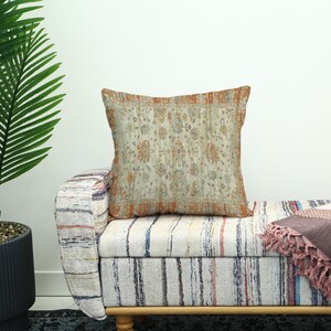 May include: A square cushion with a faded floral design in beige, orange, and blue. The cushion sits on a striped bench with a wooden frame. A potted plant is visible to the left of the cushion.