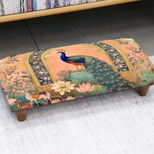 May include: A rectangular footstool with a colorful peacock design. The stool has a peach-colored background with a peacock, flowers, and other decorative elements. The legs are made of wood.