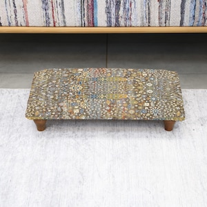 May include: A rectangular footstool with a patterned top and wooden legs. The top features a detailed design with various colors, including brown, yellow, and blue. The footstool is positioned in front of a light-colored rug and a wooden bench.