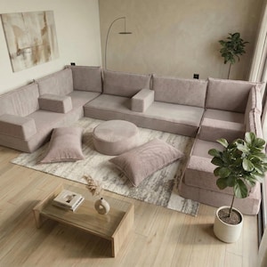 Puede incluir: Un sofá seccional modular de tela de pana rosa claro. El conjunto incluye varios cojines de asiento, reposabrazos y cojines decorativos. También hay un otomán redondo y una mesa de centro rectangular con tapa de cristal. La habitación está decorada con una alfombra y plantas.