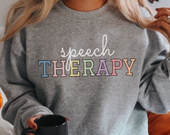 May include: A gray sweatshirt with the text "speech THERAPY" in rainbow colors. The sweatshirt is being worn by a person holding a black mug.