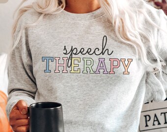 May include: A gray sweatshirt with the text "speech THERAPY" in rainbow colors. The sweatshirt is being worn by a person holding a black mug.
