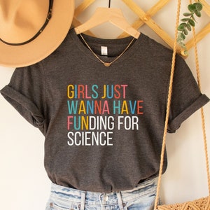 以下が含まれることがあります： カラフルな文字で「GIRLS JUST WANNA HAVE FUNDING FOR SCIENCE」と書かれたダークグレーのTシャツです。