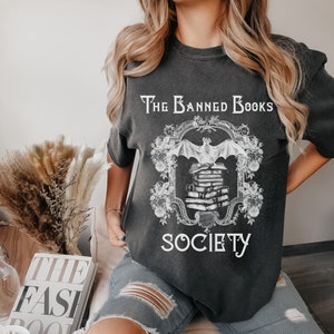 May include: A black t-shirt with a white graphic design featuring a bat and a stack of books. The text "The Banned Books Society" is printed on the shirt.