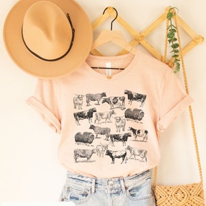 May include: A peach-colored t-shirt with a black and white illustration of various cows and bulls. The shirt is hanging on a wooden hanger with a straw hat and a woven bag.