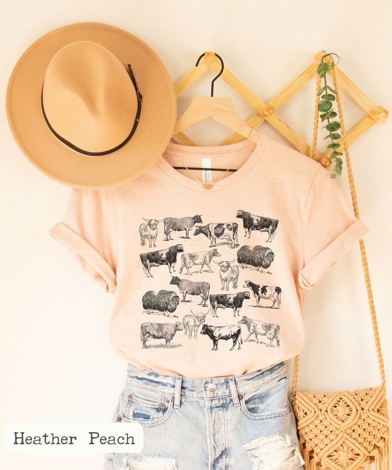 May include: A peach-colored t-shirt with a black and white illustration of various cows and bulls. The shirt is hanging on a wooden hanger with a straw hat and a woven bag.